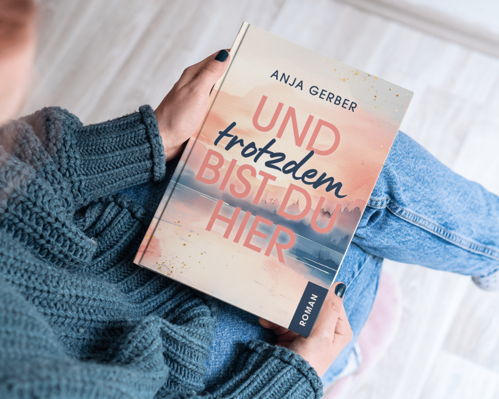 Eine Person in einem blauen Strickpullover hält ein Buch mit dem Titel „Und trotzdem bist du hier…“ von Anja Gerber in den Händen.